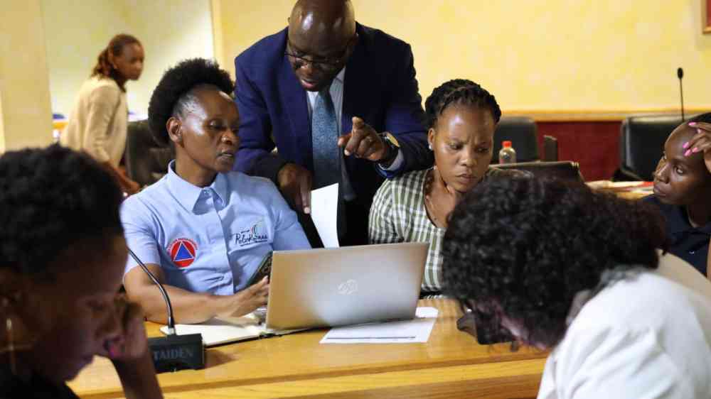 Six people are captured in the foreground of this photo. All six are engaged in collaborative work as part of a technical workshop for the Capricorn District Municipality of South Africa. The focus of the photo is on one woman, who looks at her open laptop, while speaking to a man standing behind her