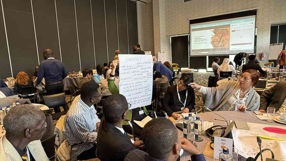 A group of people are sat down at a table, looking at a large sheet of paper to the left of the photo. The sheet of paper has been written over, and one woman motions towards the writing with a whiteboard marker, speaking to the rest of the group 