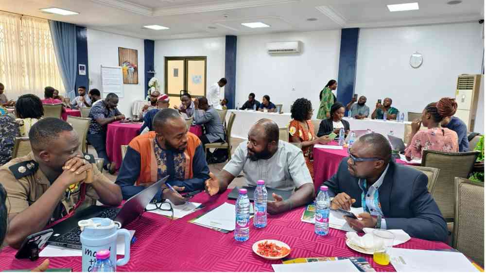 Four participants sit at a table in discussion, behind them, other groups of people are engaged in similar discussions