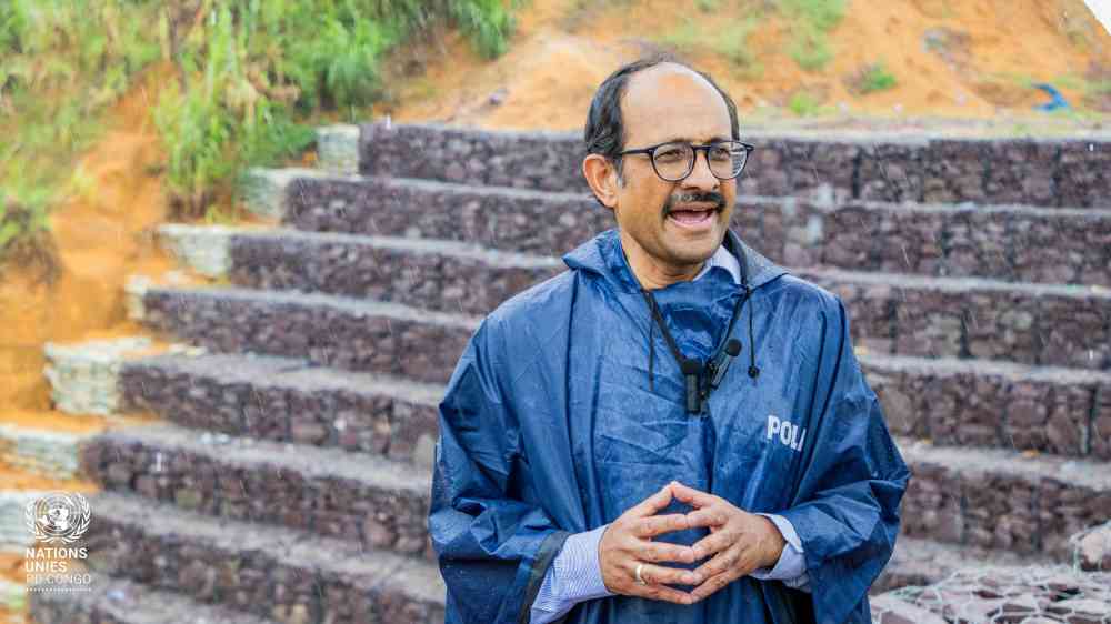 The SRSG for DRR, Kamal Kishore, stands in front of a set of stairs in an outdoor area. He is wearing a dark blue rain coat and there is a drizzle around him. He is speaking to someone who is off-camera