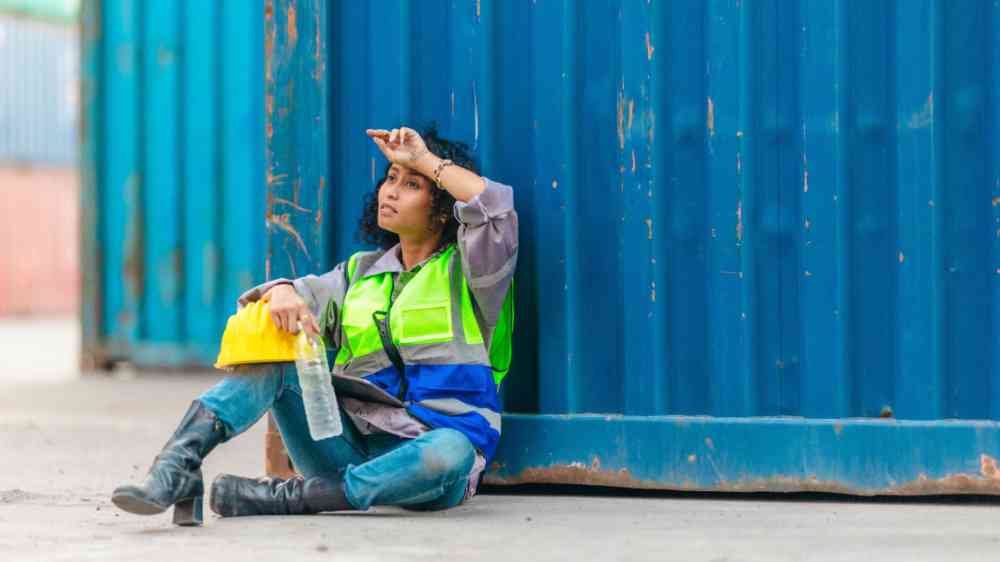 Fatigued female worker sits against shipping container, wiping sweat from her forehead while holding a water bottle and hard hat.