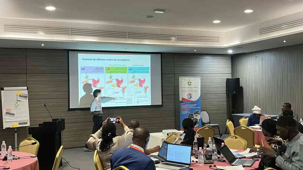 One man stands before a powerpoint presentation, which is being projected against a wall. Groups of people are sat on tables in a room, listening to the presentation