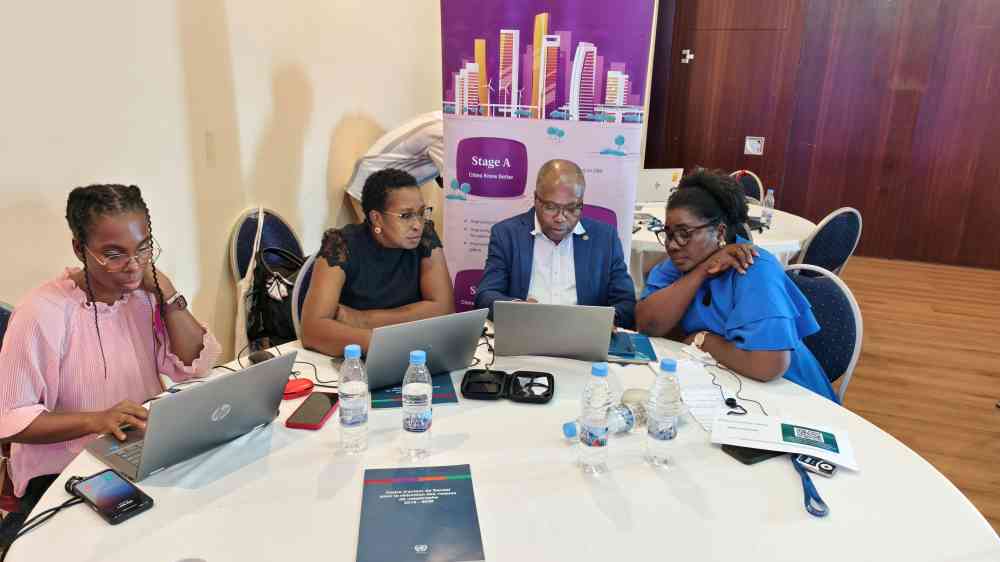 Four individuals are sat at a circular table with a white tablecloth. Three of the people have a laptops in front of them, and are reading off the screens. The last person without a laptop reaches over to read the screen of the person sat next to them. A purple Making Cities Resilient 2030 banner is placed behind them